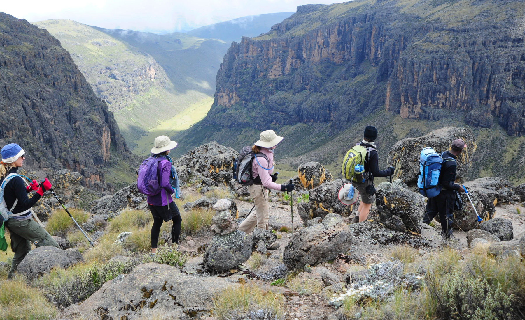 A group of hikers within Mount Kenya region. Hiking along the edge of the mountain, overlooking a valley on a partly cloudy day. 