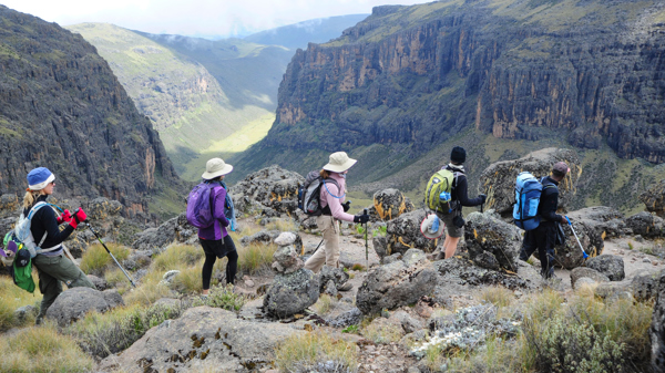 A group of hikers within Mount Kenya region. Hiking along the edge of the mountain, overlooking a valley on a partly cloudy day. 
