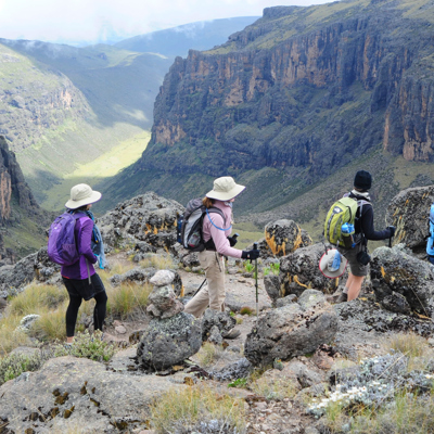 A group of hikers within Mount Kenya region. Hiking along the edge of the mountain, overlooking a valley on a partly cloudy day. 
