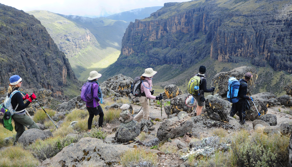 A group of hikers within Mount Kenya region. Hiking along the edge of the mountain, overlooking a valley on a partly cloudy day. 