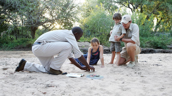 A safari guide and tracker educating young children about animal tracks within a dry river bed. Surrounded by lush, green vegetation with golden sunrays shining through the branches. 