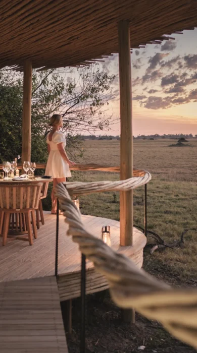 Woman stands on wooden decking watching sunset, Mombo, Moremi Game Reserve, Botswana - Yellow Zebra Safari