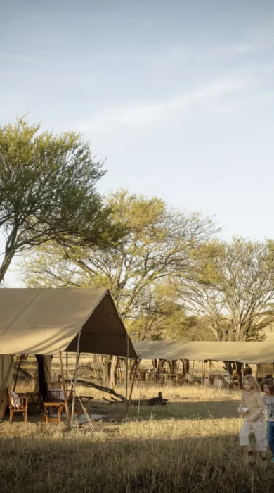 A row of tented accommodation sit in the grass as people walk by, Nomad Tanzania, Tanzania - Yellow Zebra Safari