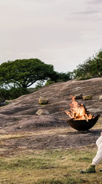 Aside a firepit, woman lounges on a camp chair in a rocky area - Yellow Zebra Safaris