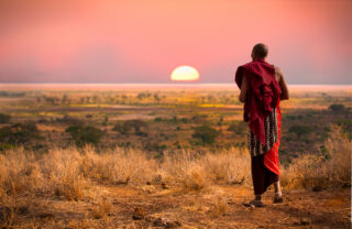 A man in traditional Masaai dress walks towards a setting sun, Mara, Kenya - Yellow Zebra Safaris