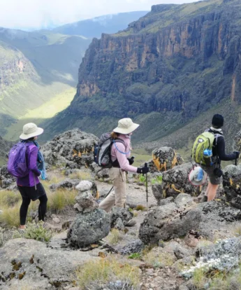 A group of hikers within Mount Kenya region. Hiking along the edge of the mountain, overlooking a valley on a partly cloudy day.